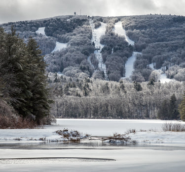 Scenic winter view of Wachusett Mountain Ski Area in Massachusetts, featuring snow-covered trails, ski lifts, and frosted trees with a frozen lake in the foreground. Ideal for skiing, snowboarding, and New England winter getaways.