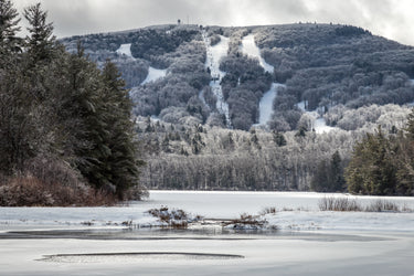 Scenic winter view of Wachusett Mountain Ski Area in Massachusetts, featuring snow-covered trails, ski lifts, and frosted trees with a frozen lake in the foreground. Ideal for skiing, snowboarding, and New England winter getaways.